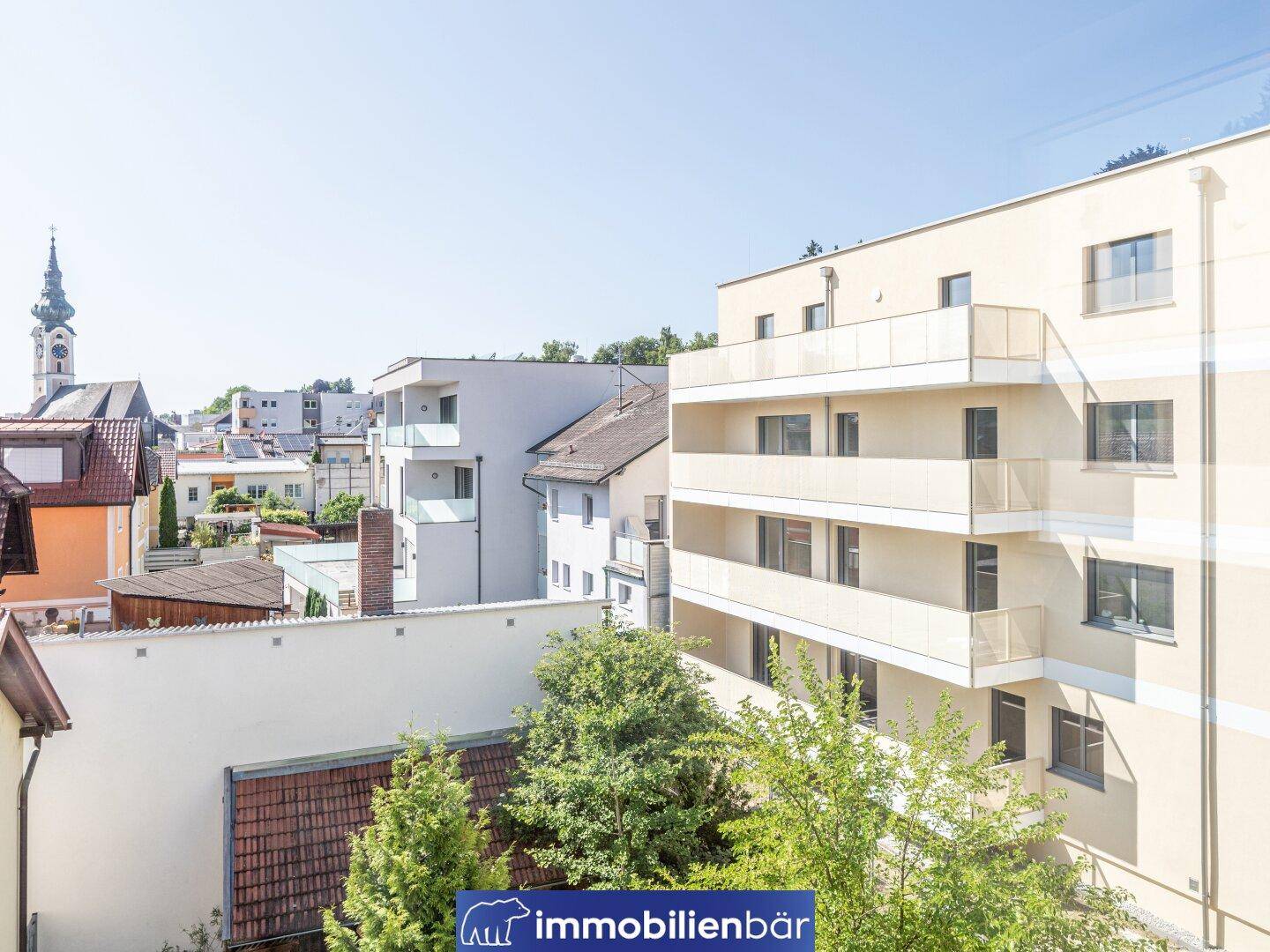 Blick auf die Balkone des Neubaus und die angrenzende Wohnbebauung mit viel Grün und einem Kirchturm.