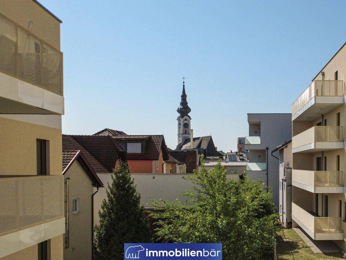 Blick aus einem Fenster oder Balkon auf einen Kirchturm und die umliegende Stadtbebauung mit Bäumen.