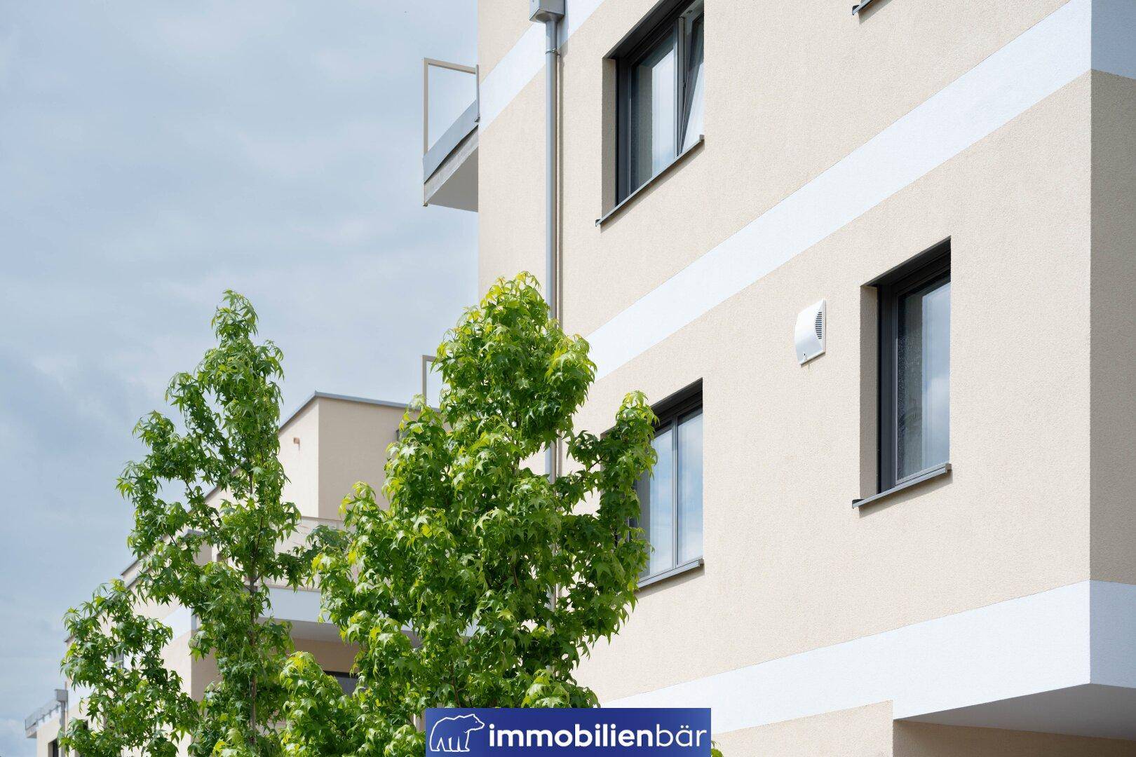 Detailansicht der modernen Fassade des Neubaus mit Fenstern und einem kleinen Balkon, umgeben von Bäumen.
