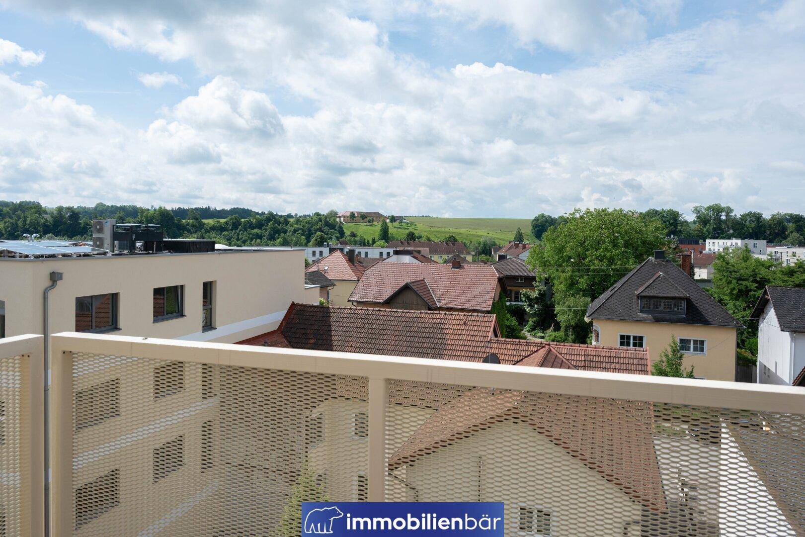 Blick von einem Balkon über Dächer und die umliegende grüne Landschaft unter bewölktem Himmel.