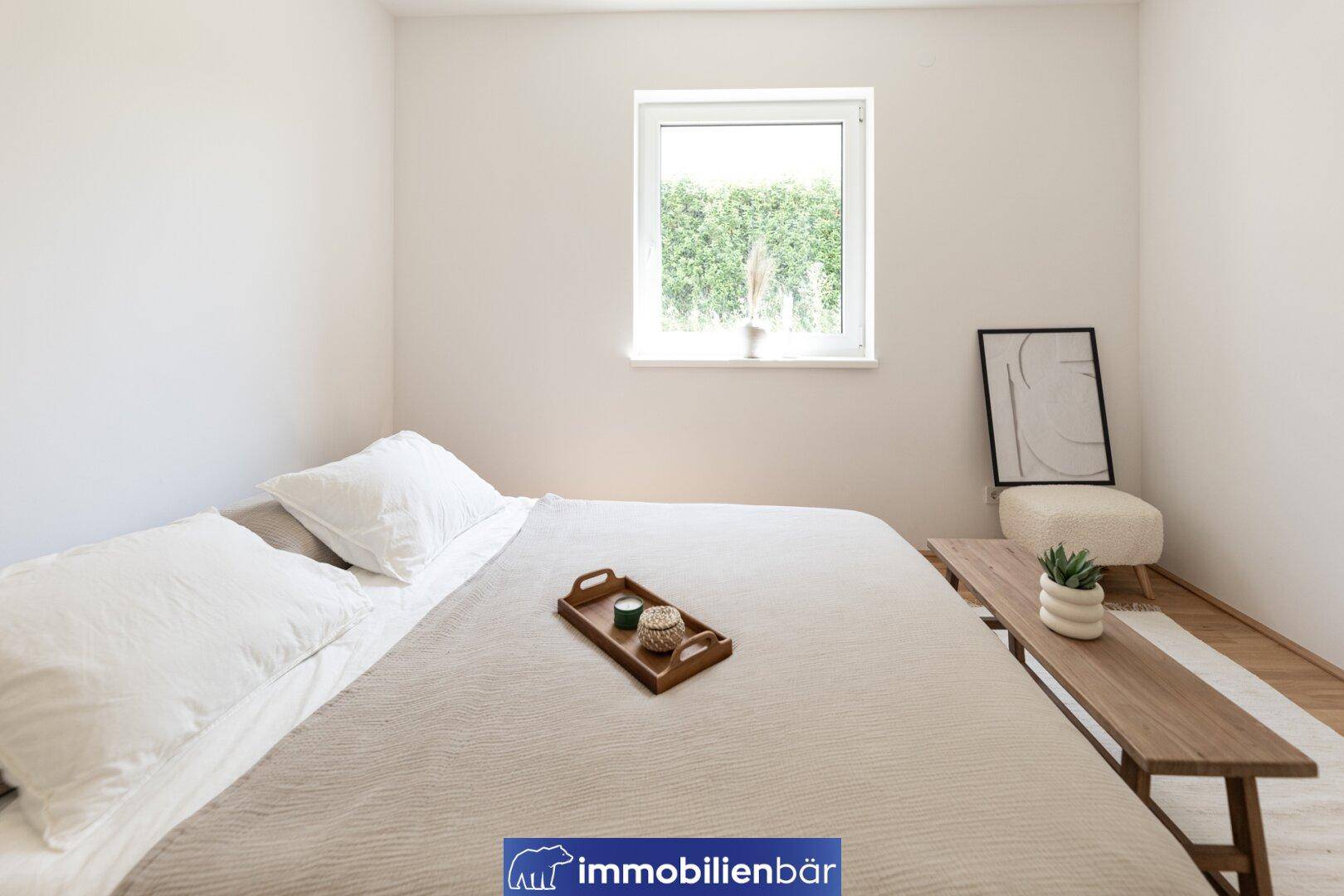 Gemütliches Schlafzimmer mit beigem Bett und Blick durch das Fenster auf eine grüne Hecke.