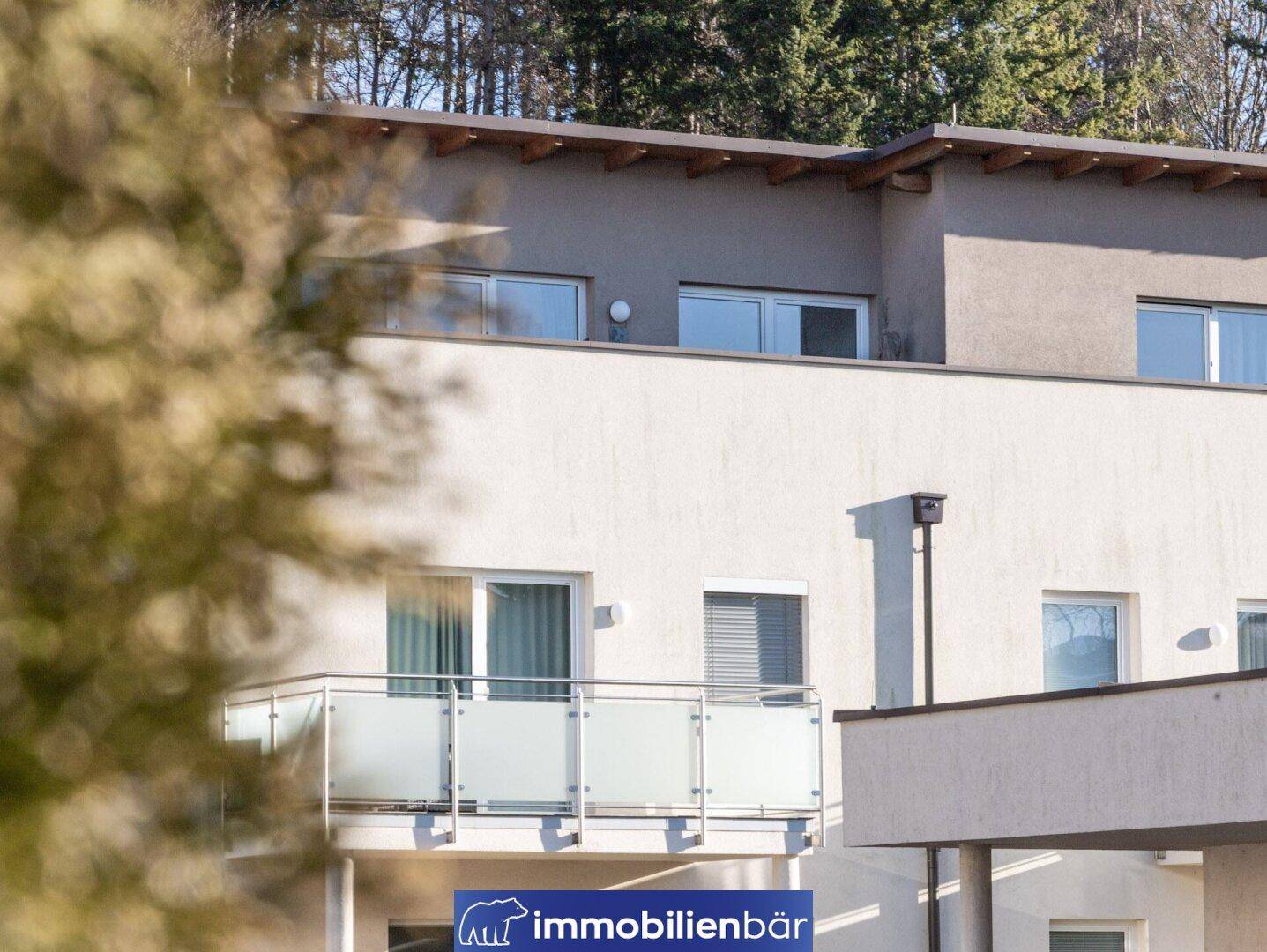 Nahaufnahme der Gebäudefassade mit einem Balkon und modernen Fenstern, umgeben von Bäumen.