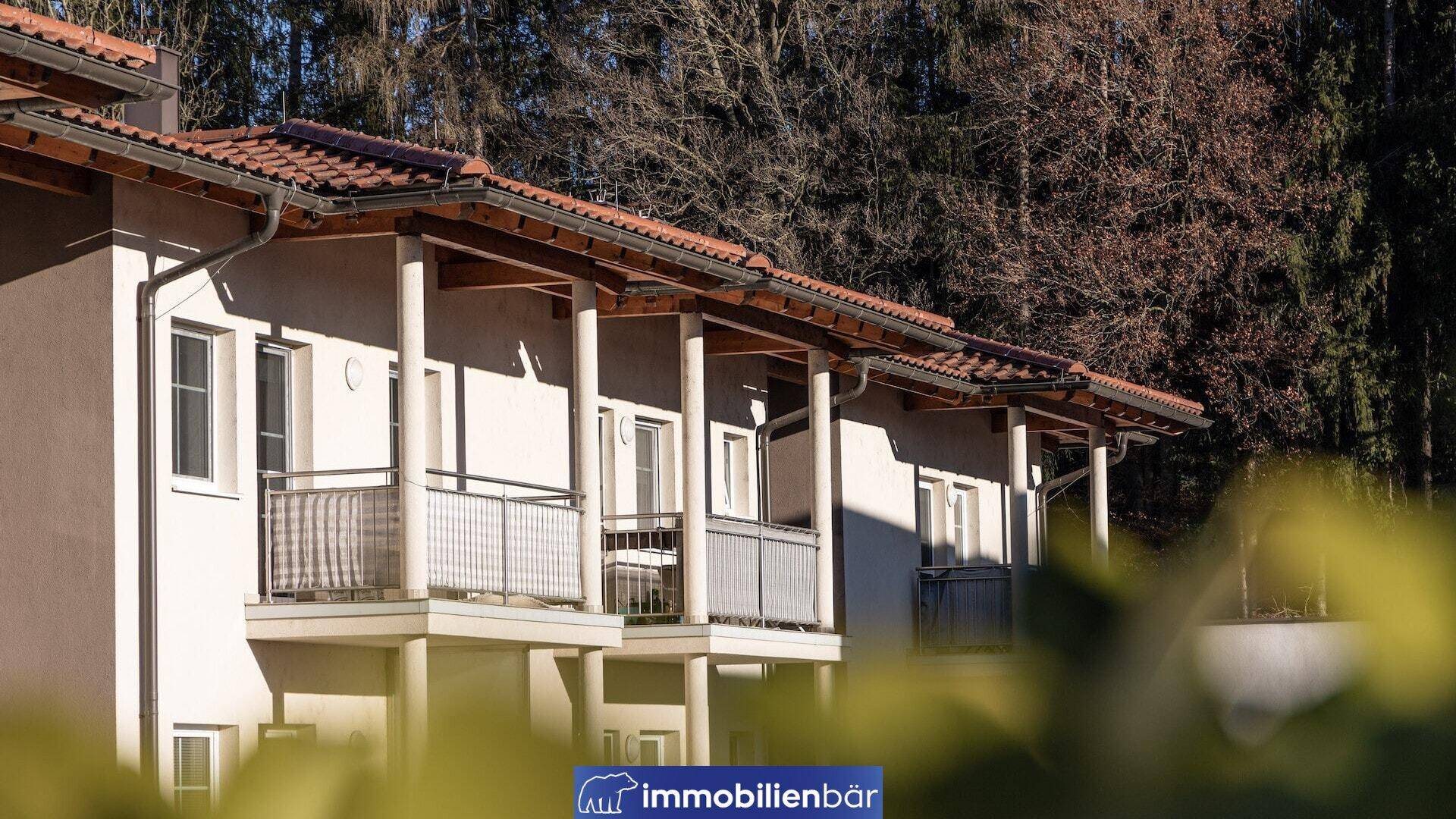 Reihe von Wohneinheiten mit überdachten Balkonen und traditionellen Ziegeldächern vor einem Waldhintergrund.