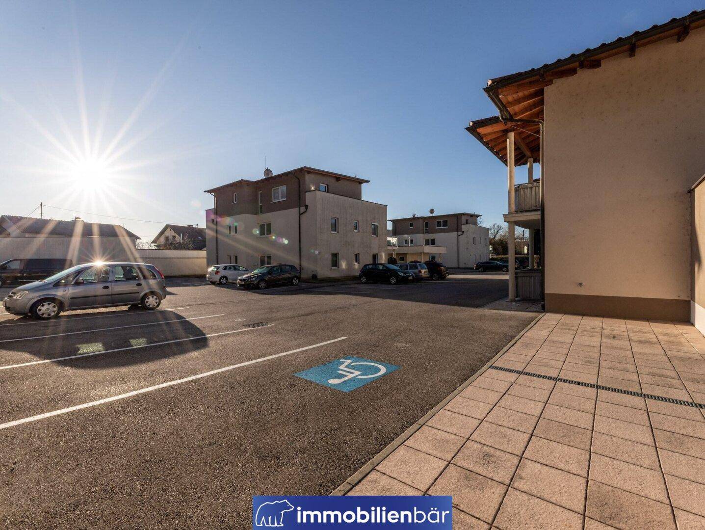 Außenansicht mit Parkplätzen, darunter ein Behindertenparkplatz, und Wohngebäuden im sonnigen Hintergrund.