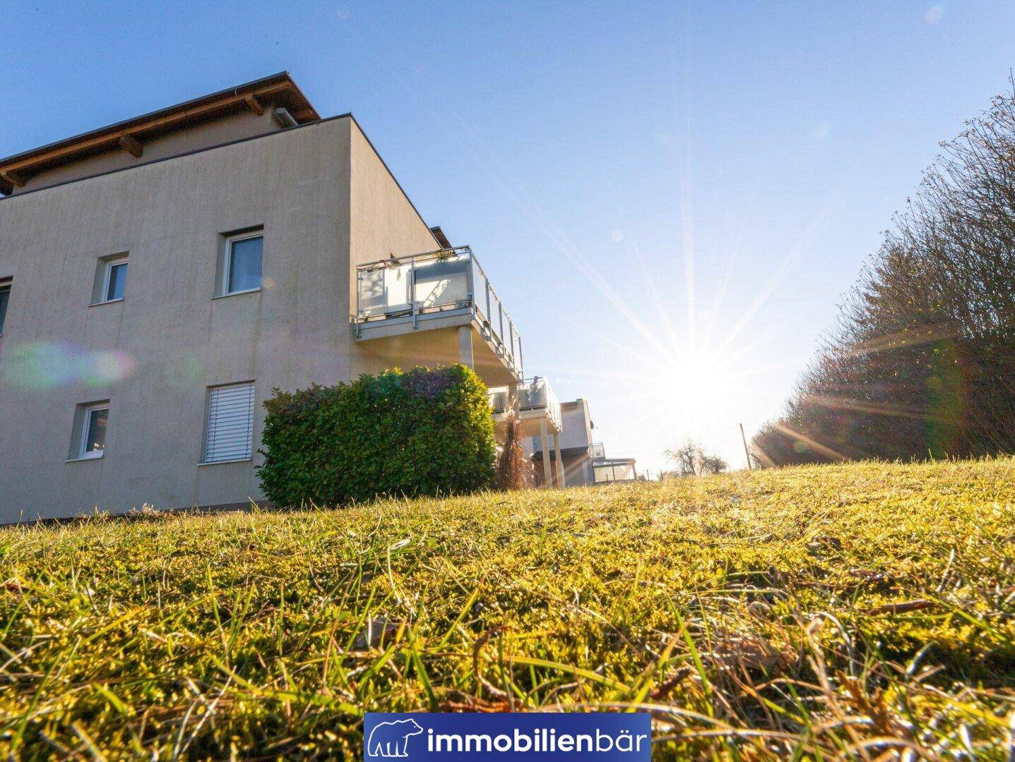 Modernes Wohngebäude mit Balkonen, aufgenommen von einer sonnigen, grasbewachsenen Anhöhe.