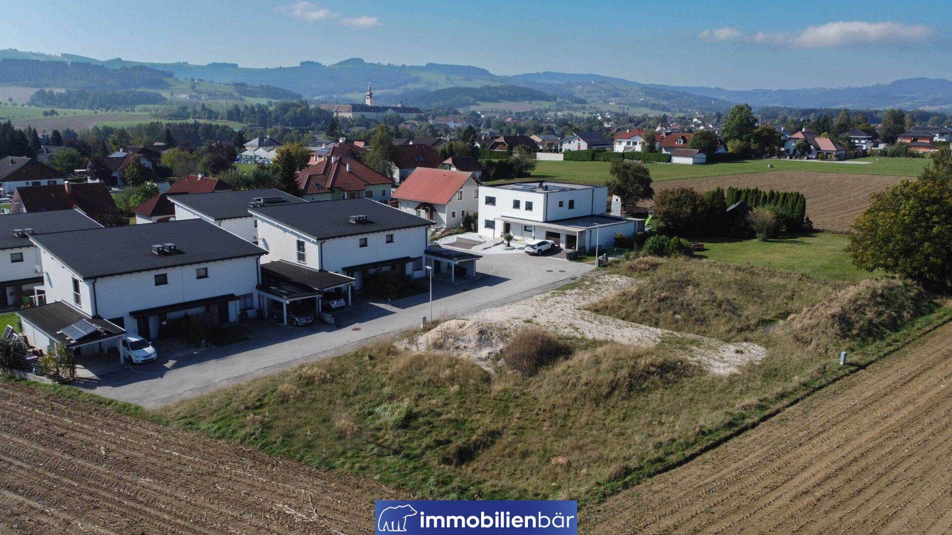 Weitläufige Luftaufnahme eines Baugrundstücks mit angrenzenden Häusern und einem Dorf in der Ferne.