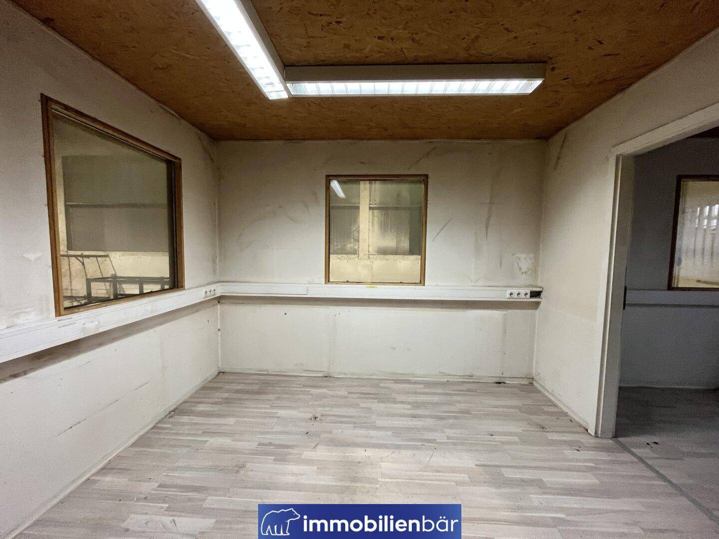 Kleines Büro mit Holzdecke, Laminatboden und einem Fenster zur angrenzenden Halle.