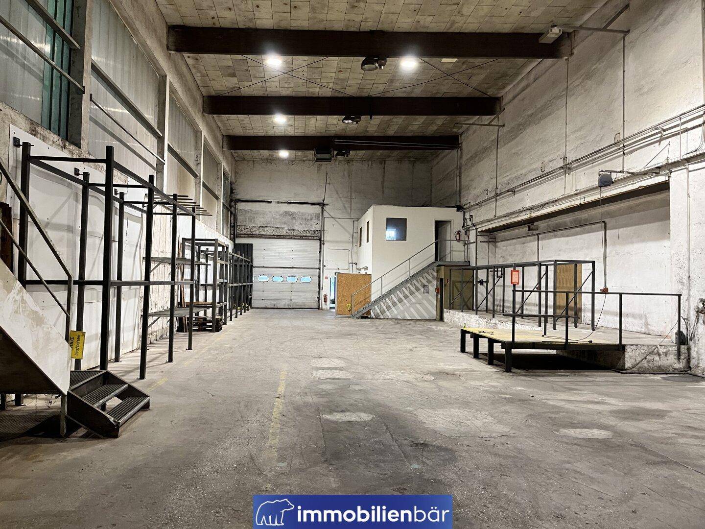 Geräumige Lagerhalle mit Betonboden, Rolltor und einem kleinen Bürogebäude im hinteren Bereich.