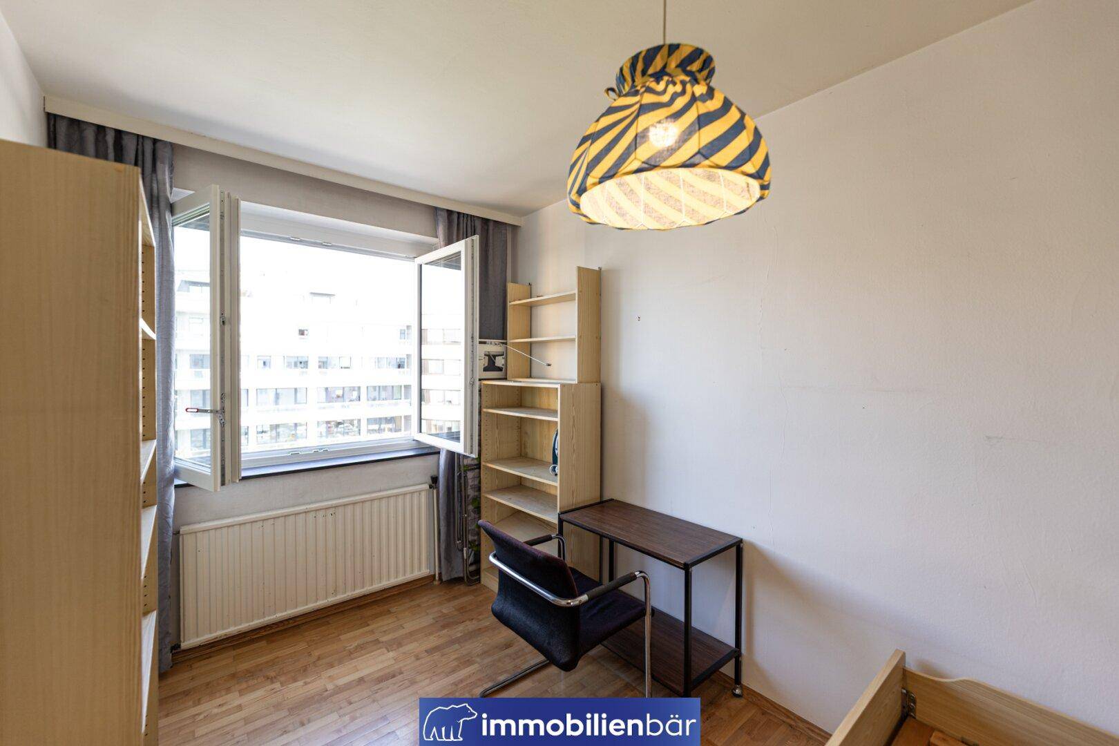 Helles Schlafzimmer mit Parkettboden, großem Fenster und einfacher Einrichtung.