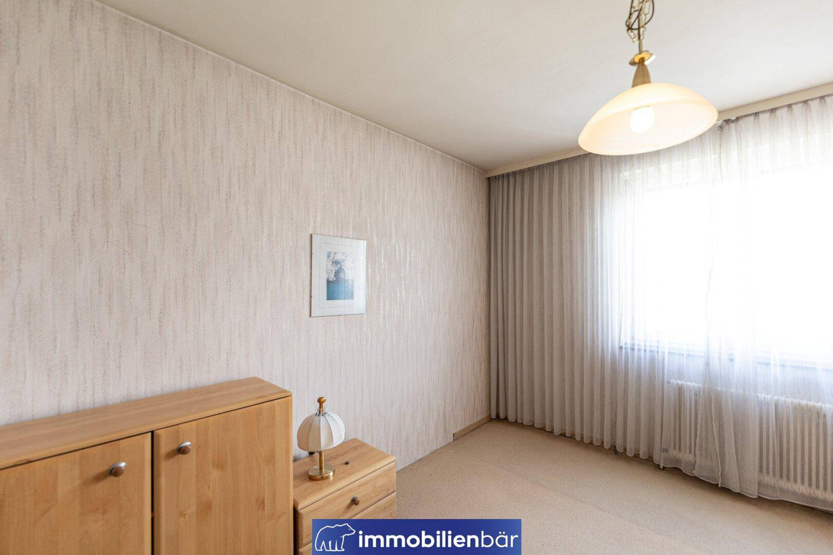 Ruhiges Schlafzimmer mit Teppichboden, hellem Fenster und einfacher Möblierung.