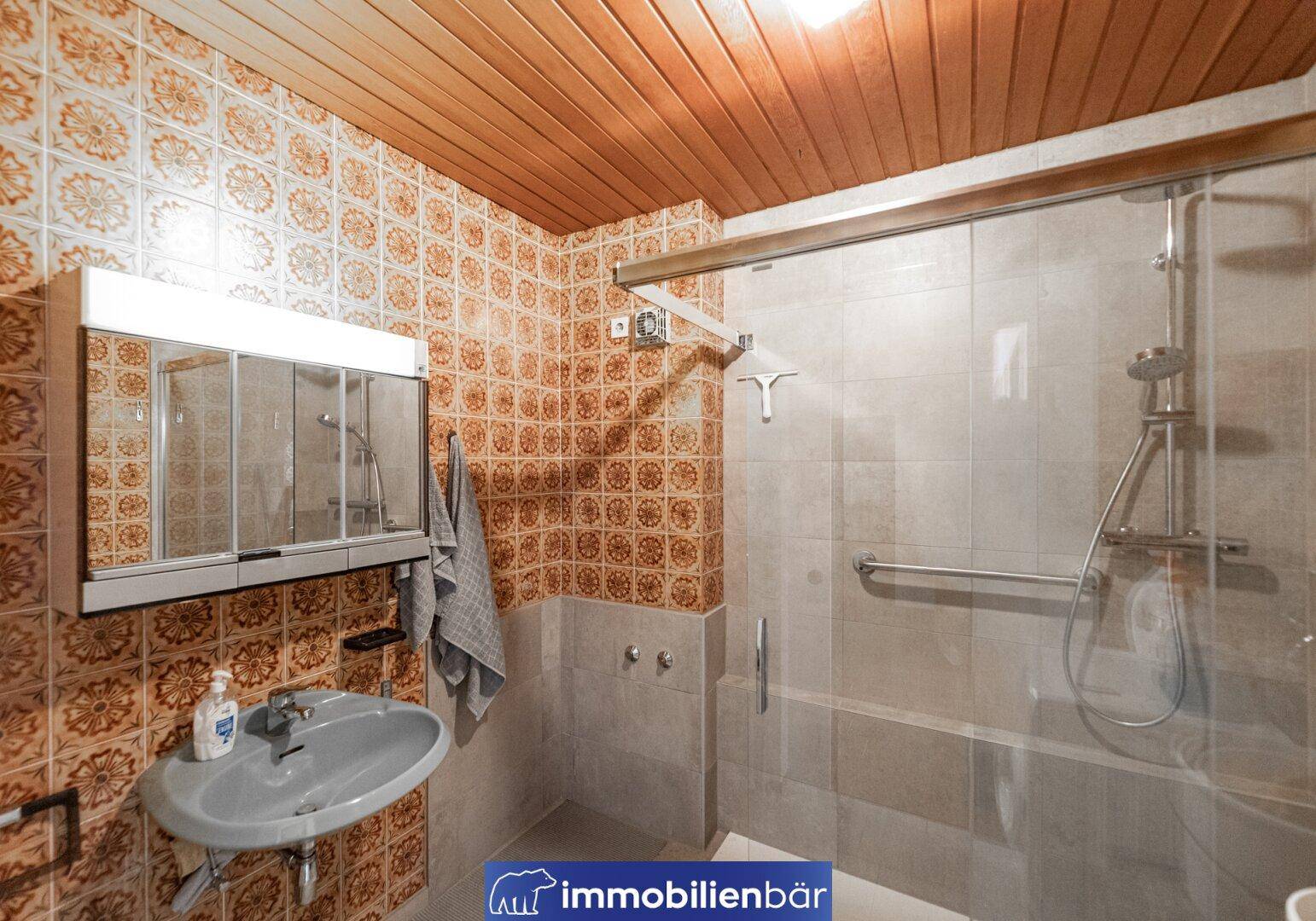 Badezimmer im 80er-Jahre-Stil mit Dusche, Waschbecken und gemusterten Wandfliesen.