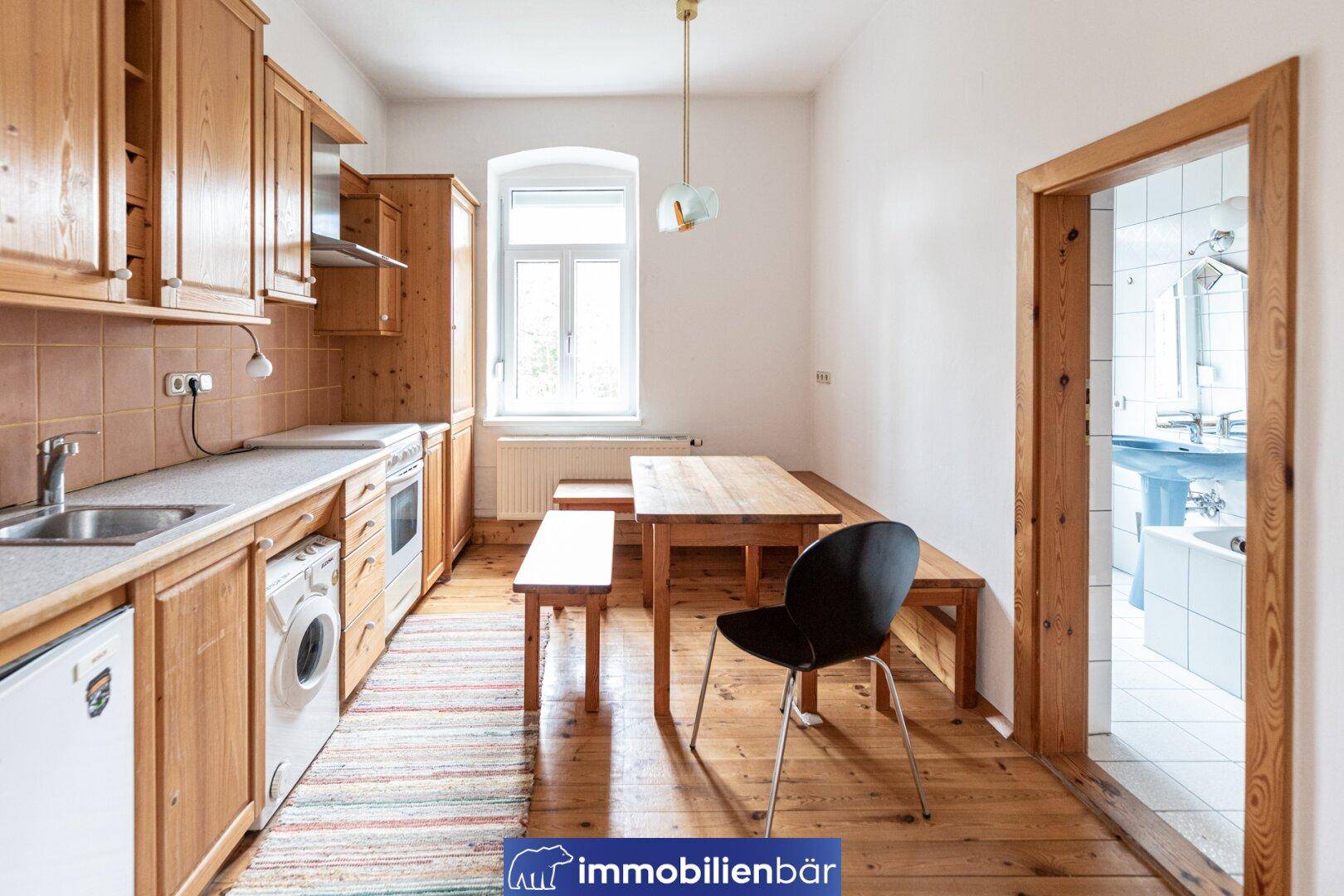 Helle Wohnküche mit Holzmöbeln, Essbereich und Blick in ein gefliestes Badezimmer.