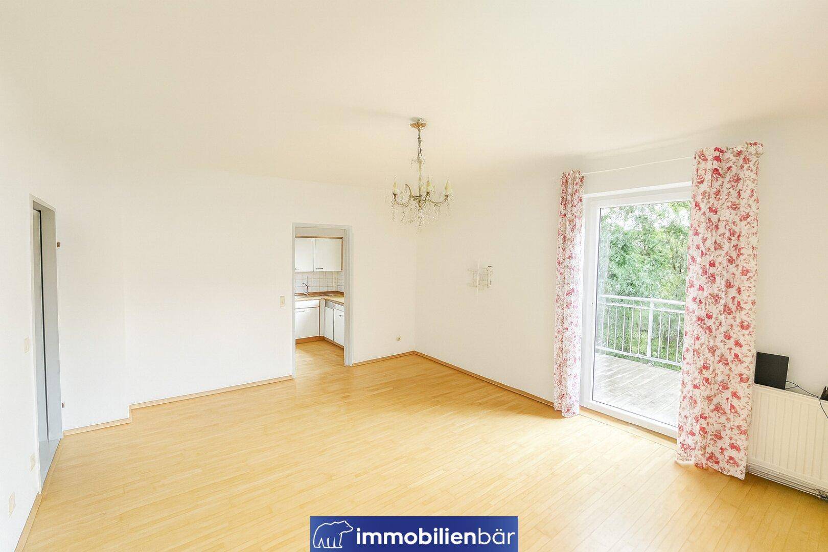 Geräumiges Wohnzimmer mit hellem Laminatboden, Balkonzugang und Blick in die separate Küche.