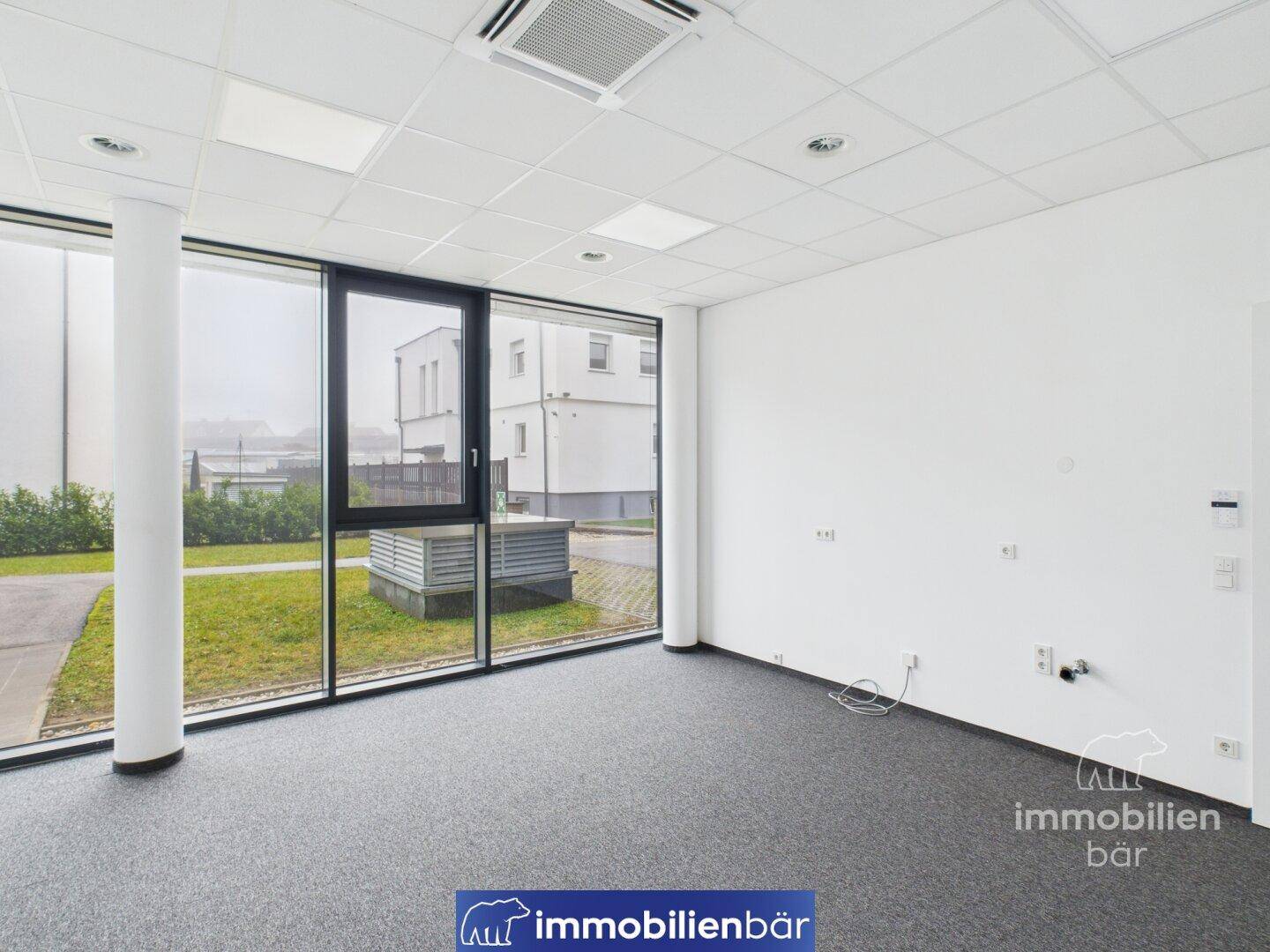 Offener Bürobereich mit Teppichboden und großen Fenstern, die einen Blick auf den Außenbereich ermöglichen und eine luftige Atmosphäre bieten.