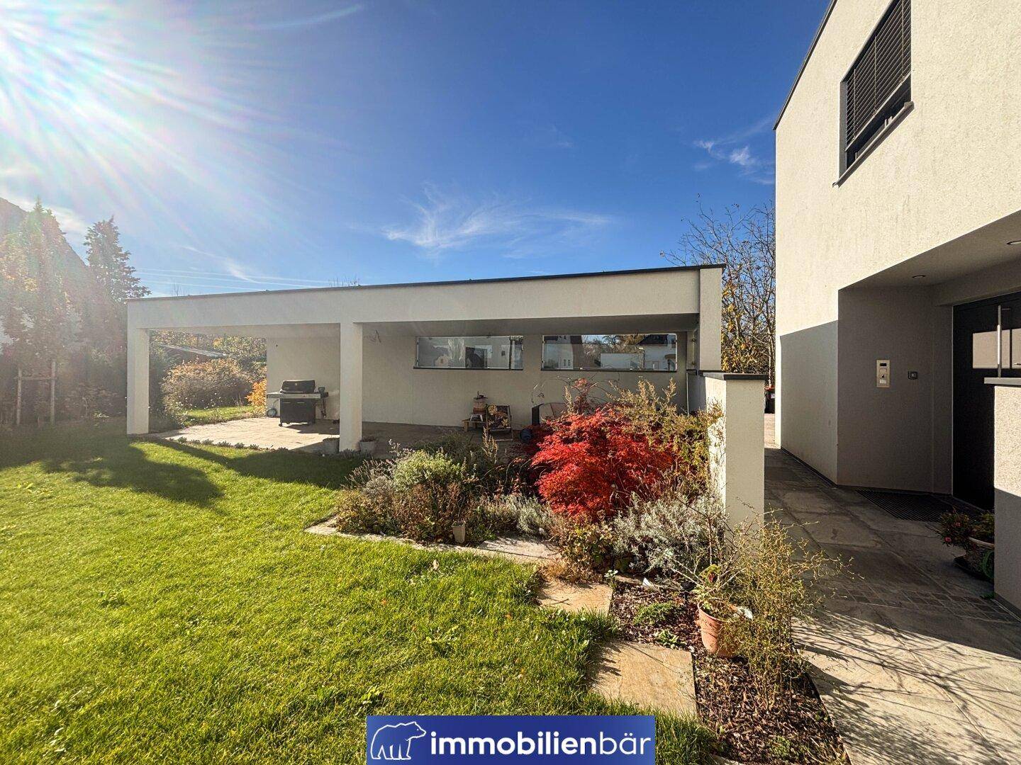 Sonniger Garten mit Rasenfläche, Pflanzen und einem modernen Carport neben dem Haus.