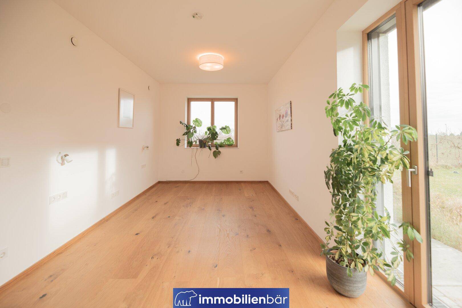 Geräumiges Zimmer mit Holzboden und großen Fenstern, die viel Tageslicht und Blick ins Grüne bieten.