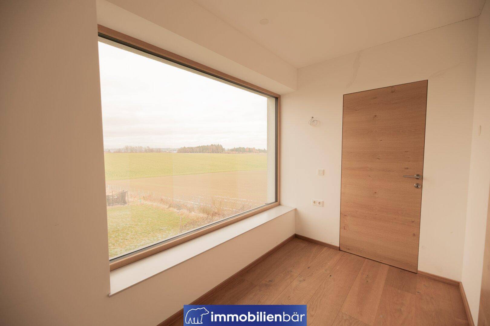 Geräumiges Zimmer mit Holzboden und Panoramafenster, das einen weiten Blick in die Natur ermöglicht.