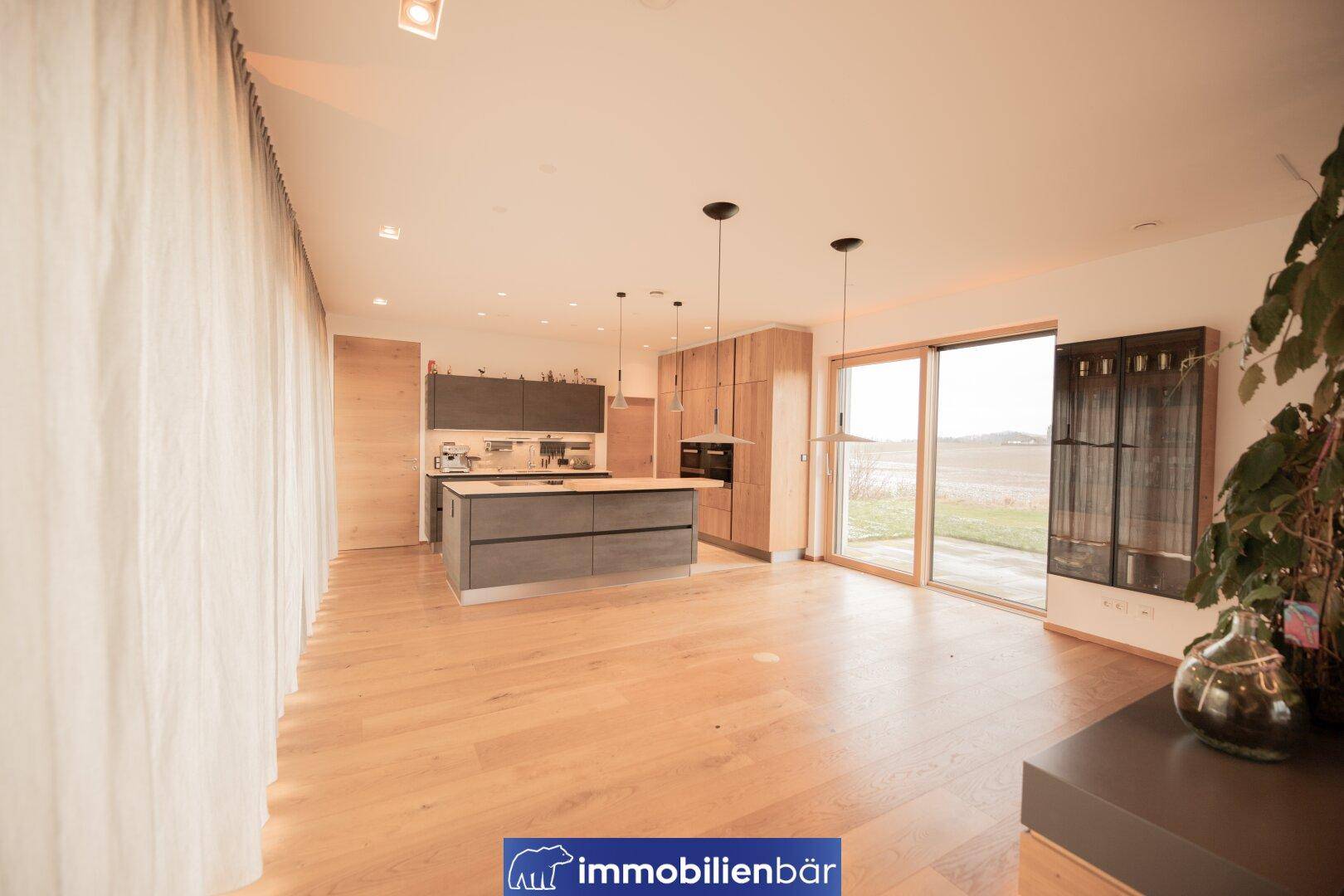 Geräumiger offener Wohnbereich mit moderner Küche, Holzboden und Zugang zur Terrasse mit Blick ins Grüne.