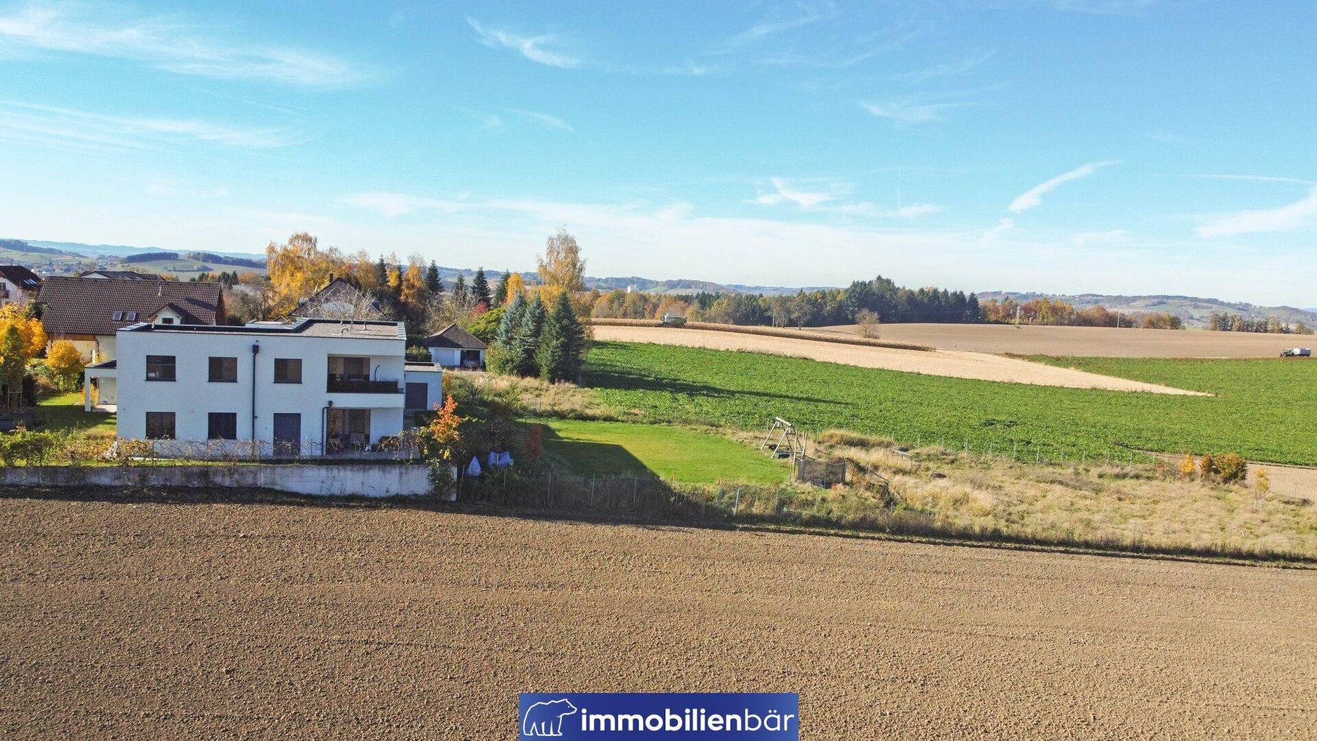 Weitläufige Landschaft mit Feldern und einem modernen Wohnhaus in der Ferne unter blauem Himmel.