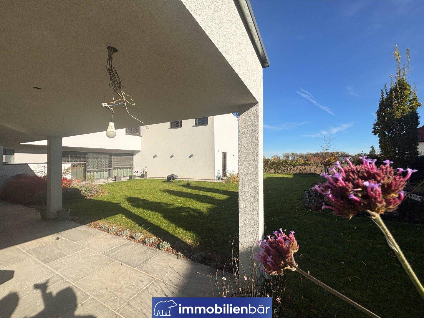 Überdachte Terrasse mit Fliesenboden und Blick auf den grünen Garten und das moderne Gebäude.