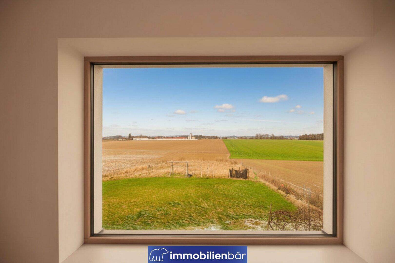 Weitläufiger Blick aus dem Fenster auf Felder und grüne Wiesen unter blauem Himmel.