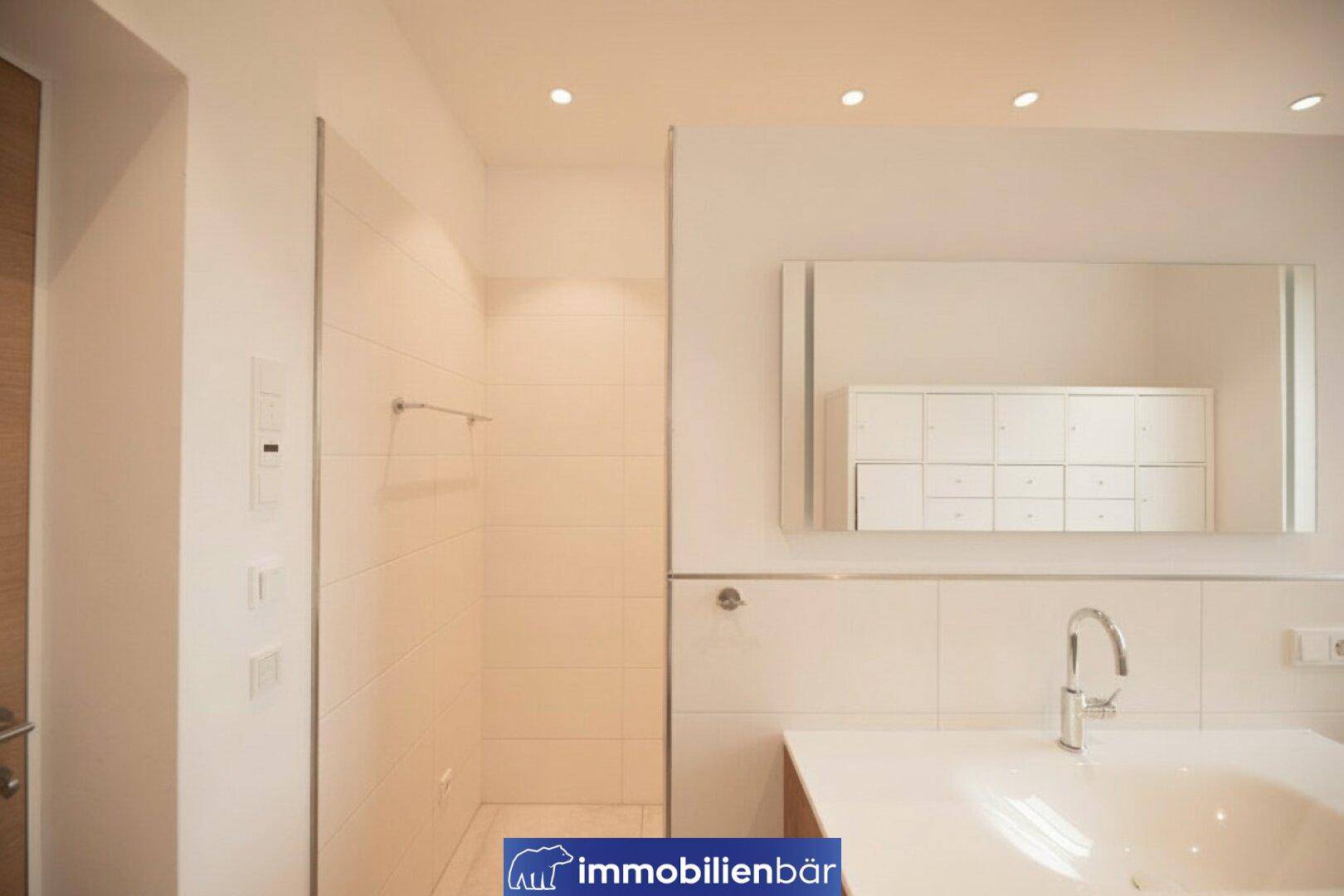 Modernes Badezimmer mit begehbarer Dusche, Waschtisch und großem Spiegel, in hellen Tönen gehalten.