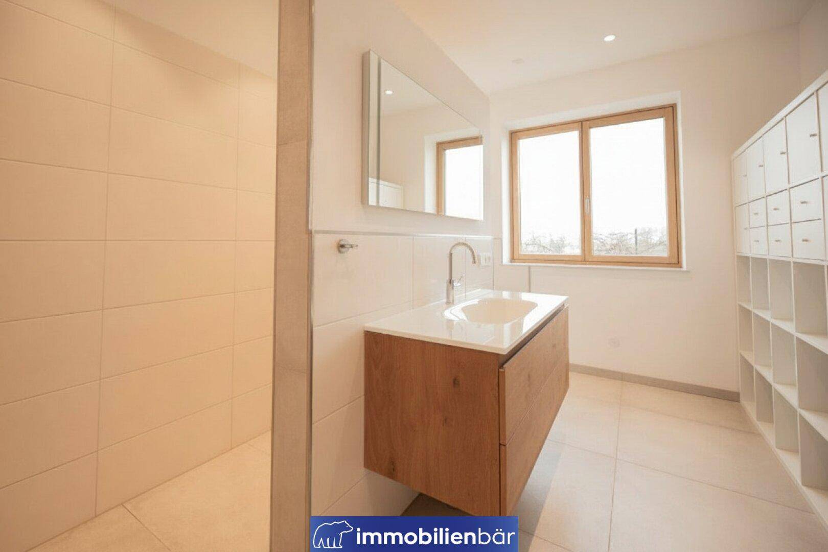 Modernes Badezimmer mit Waschtisch, großem Spiegel und begehbarer Dusche, hell gefliest.