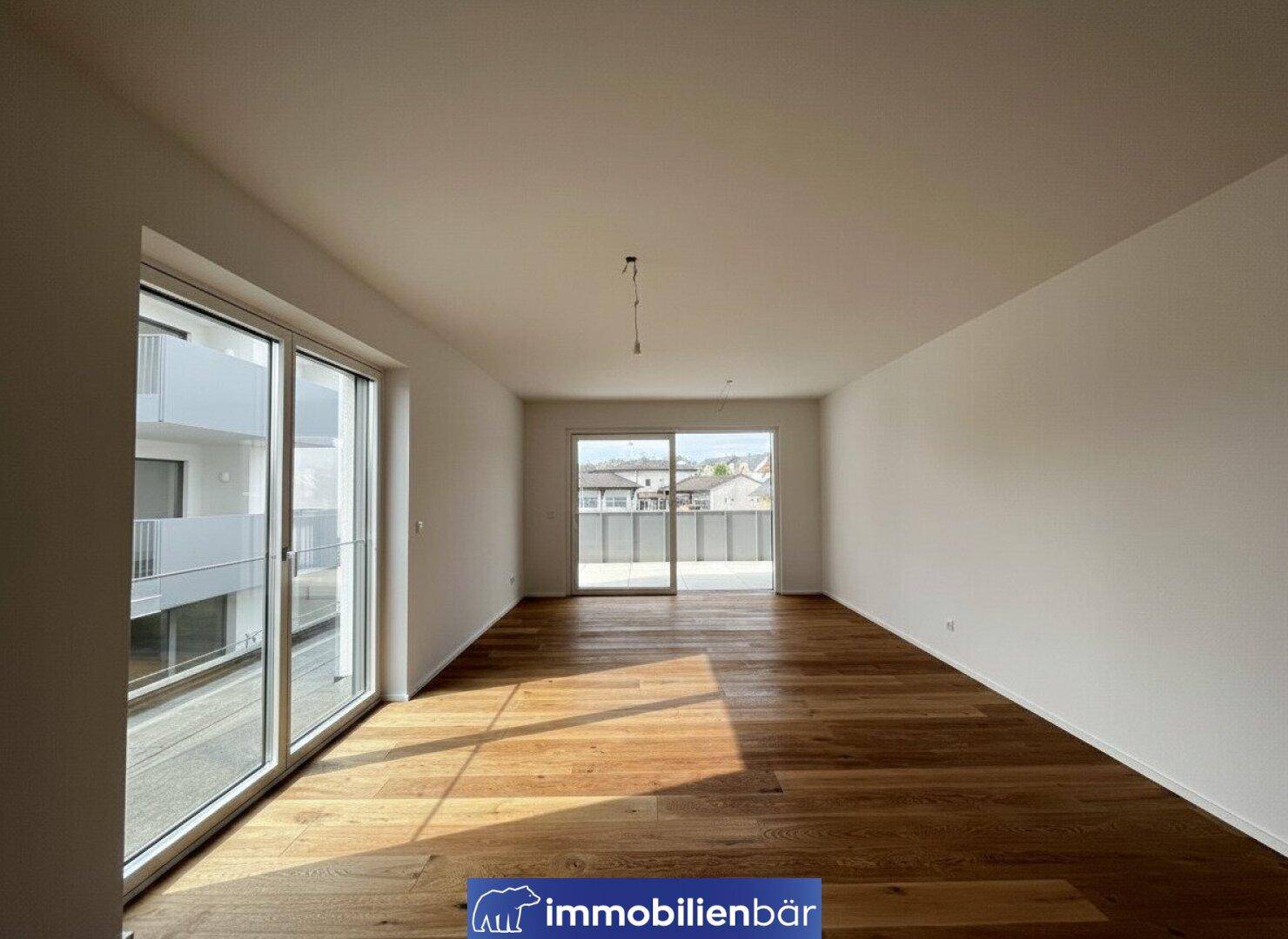 Geräumiges, leeres Wohnzimmer mit Holzboden und großen Fenstern, die viel Tageslicht hereinlassen.