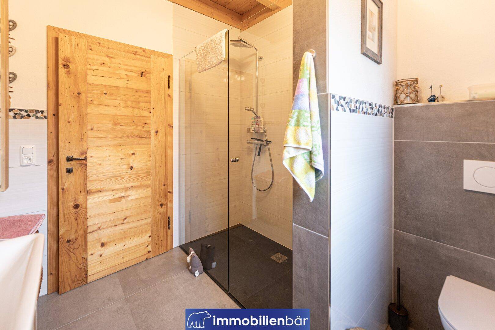 Modernes Badezimmer mit begehbarer Dusche, grauen Fliesen und Holztür.