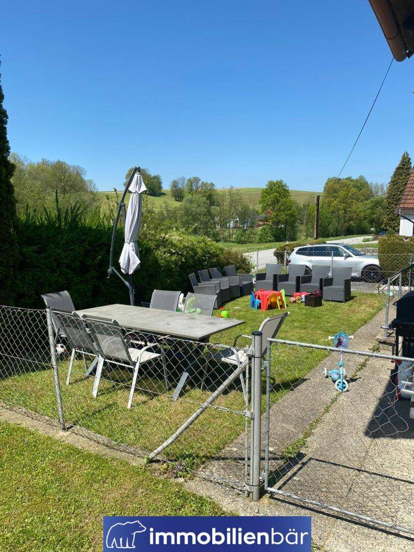 Grüner Garten mit möblierter Terrasse und Spielzeug unter strahlend blauem Himmel.