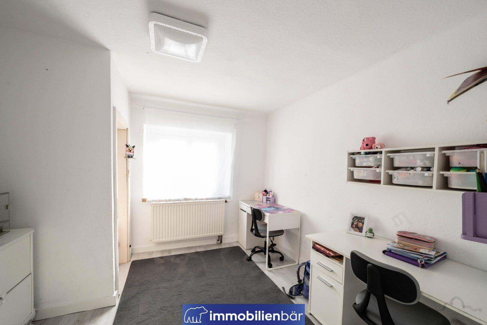 Helles Kinderzimmer mit zwei Schreibtischen, Stühlen und einem Fenster mit Heizkörper.