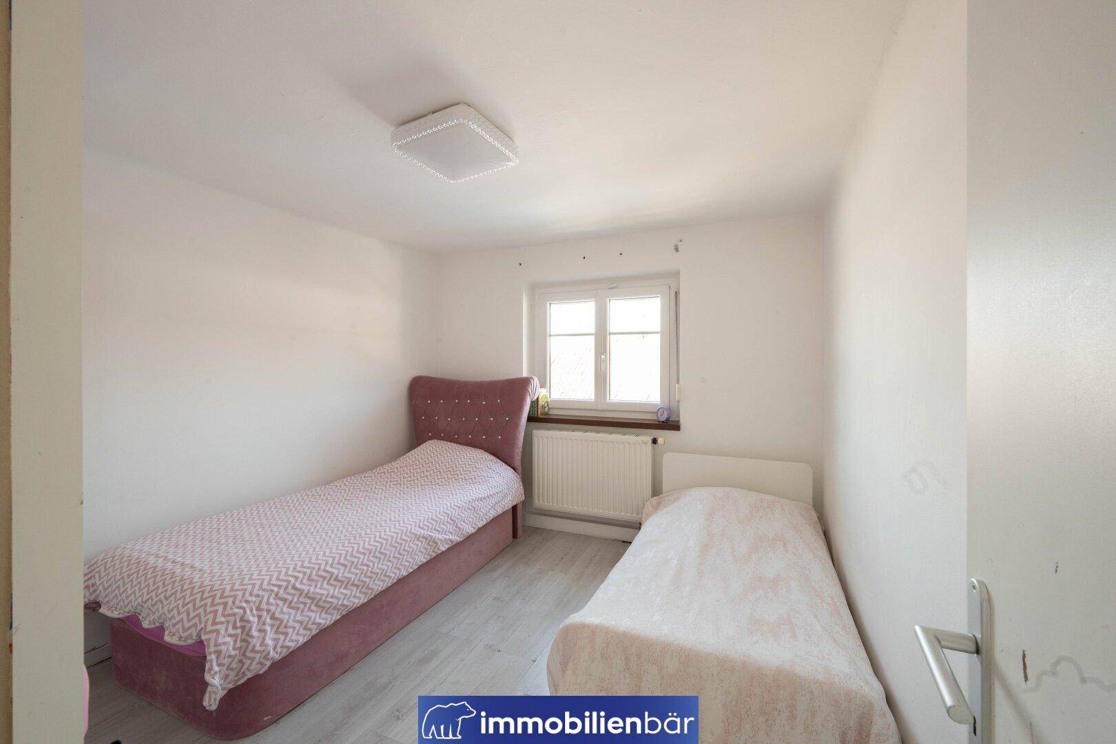 Kinderzimmer mit zwei Einzelbetten, Fenster und Heizkörper, in hellen Farben.
