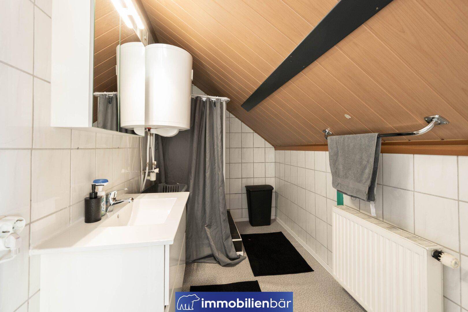 Badezimmer mit Dusche, Waschbecken und Dachschräge, in hellen Fliesen gehalten.