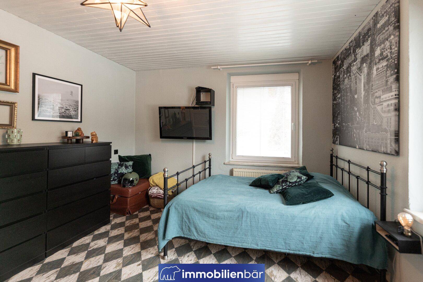 Schlafzimmer mit Metallbett, dunkler Kommode und einem auffälligen schwarz-weiß gemusterten Boden.