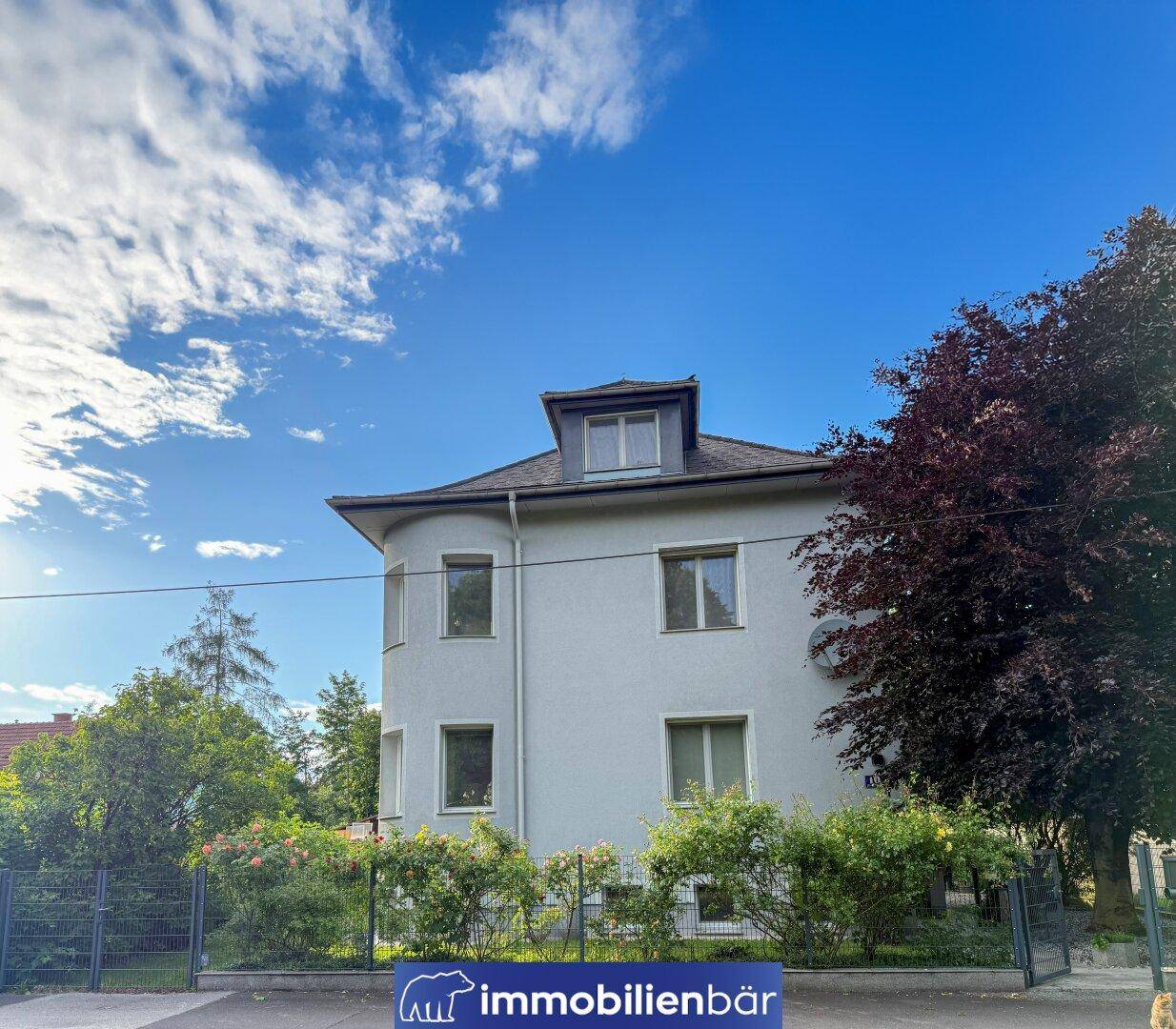 Klassisches Gebäude mit heller Fassade, vielen Fenstern und umzäuntem Vorgarten.