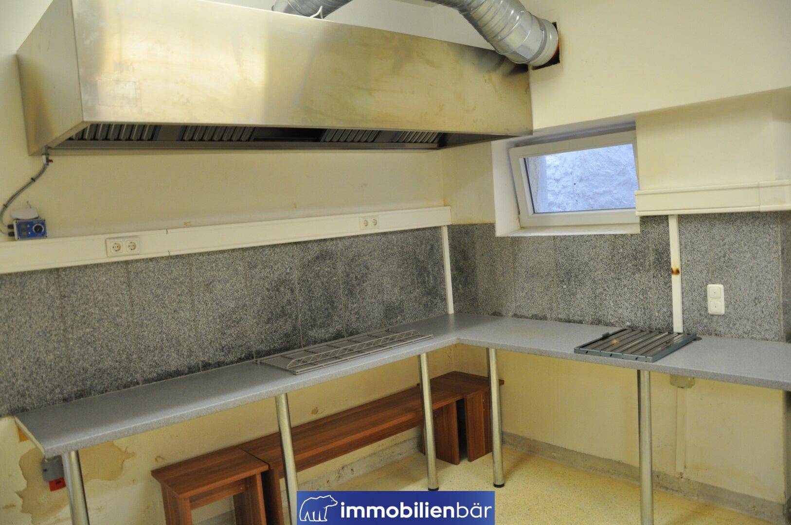 Gewerbeküche mit großer Dunstabzugshaube, langen Arbeitsflächen und einem Fenster, ideal für Restaurantbetrieb.