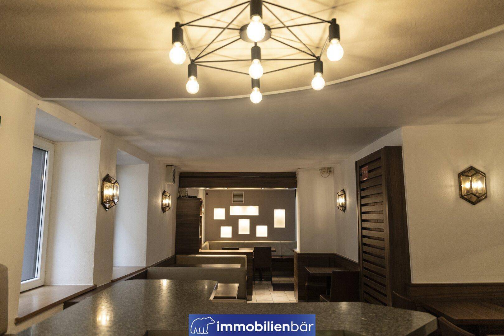 Blick über eine Theke in den Gastraum mit Sitzbänken, Holztischen und einer modernen Deckenleuchte.