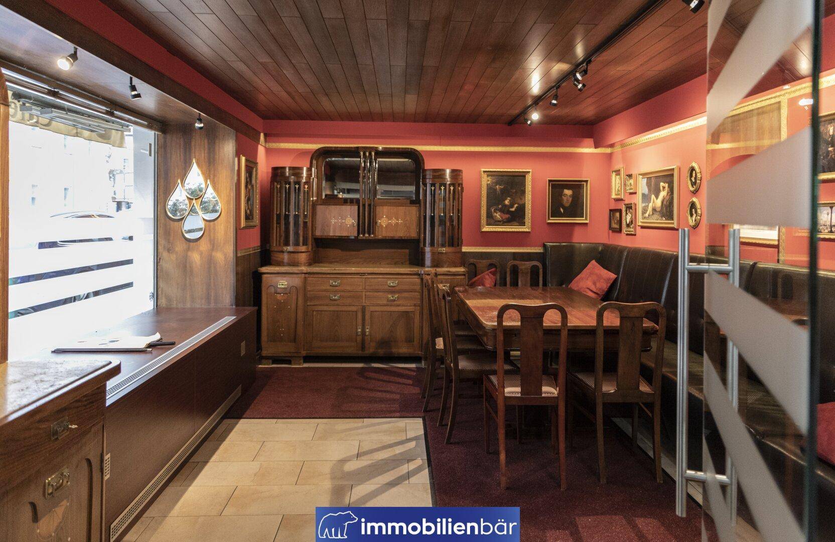 Traditioneller Gastraum mit dunklen Holzmöbeln, roter Wandfarbe und Deckenpaneelen, dekoriert mit Gemälden.