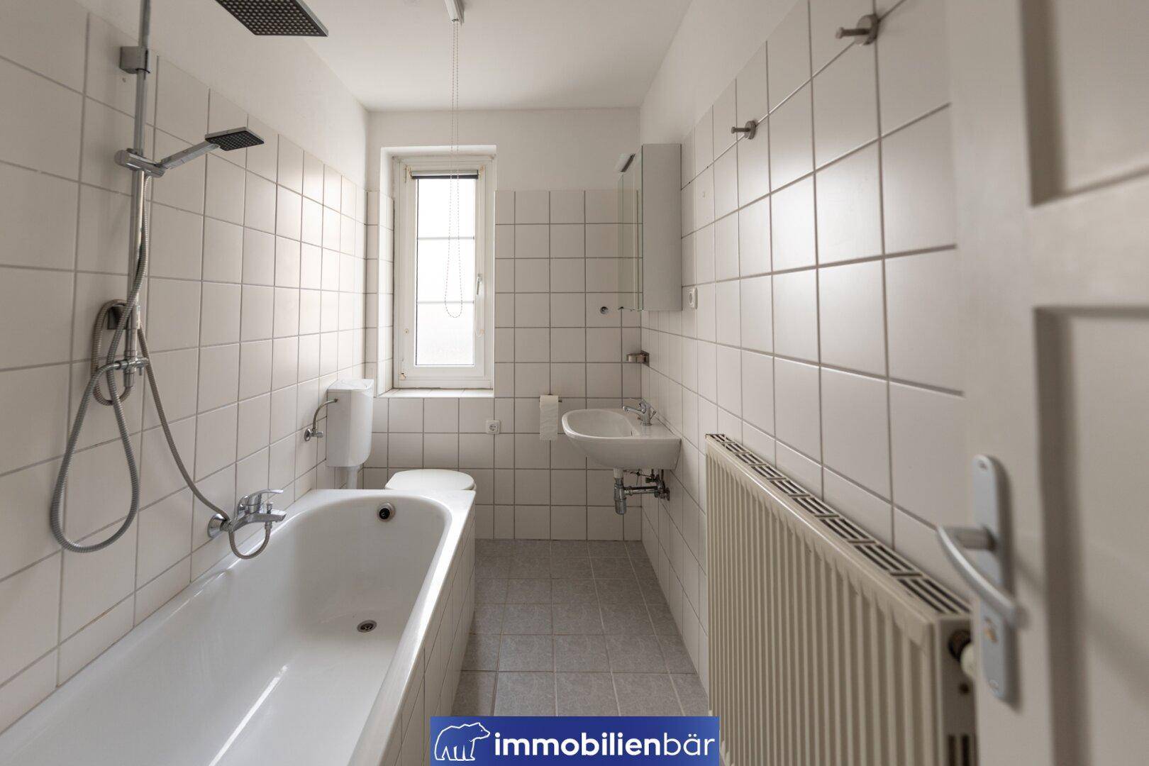 Funktionales Badezimmer mit Badewanne, Dusche und Fenster, komplett gefliest in Weiß.