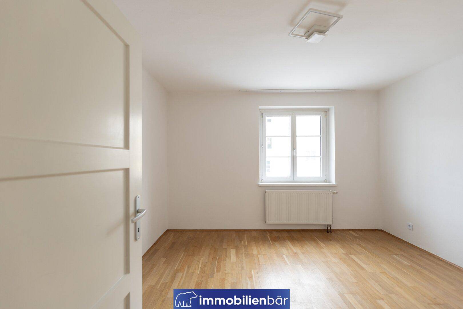 Helles, leeres Zimmer mit Holzboden und großem Fenster, bereit zur individuellen Gestaltung.