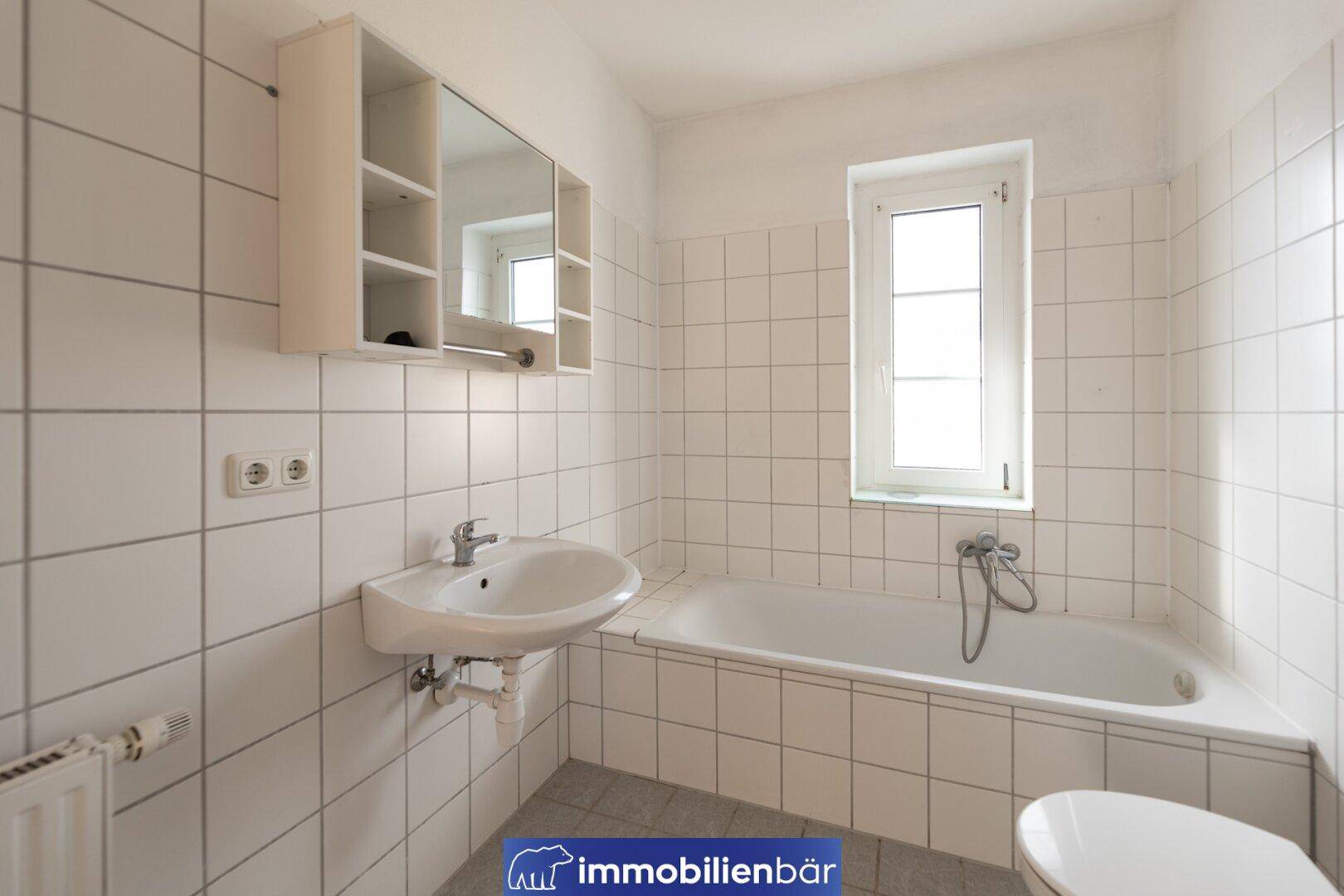 Klassisches Badezimmer mit Badewanne, Waschbecken und Fenster, vollständig gefliest in Weiß.