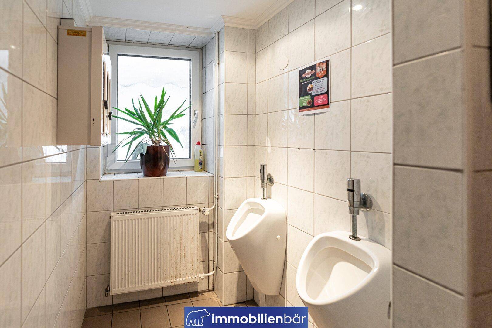 Herren-Gästetoilette mit zwei Urinalen, Fliesenwänden und einem Fenster für Belüftung.