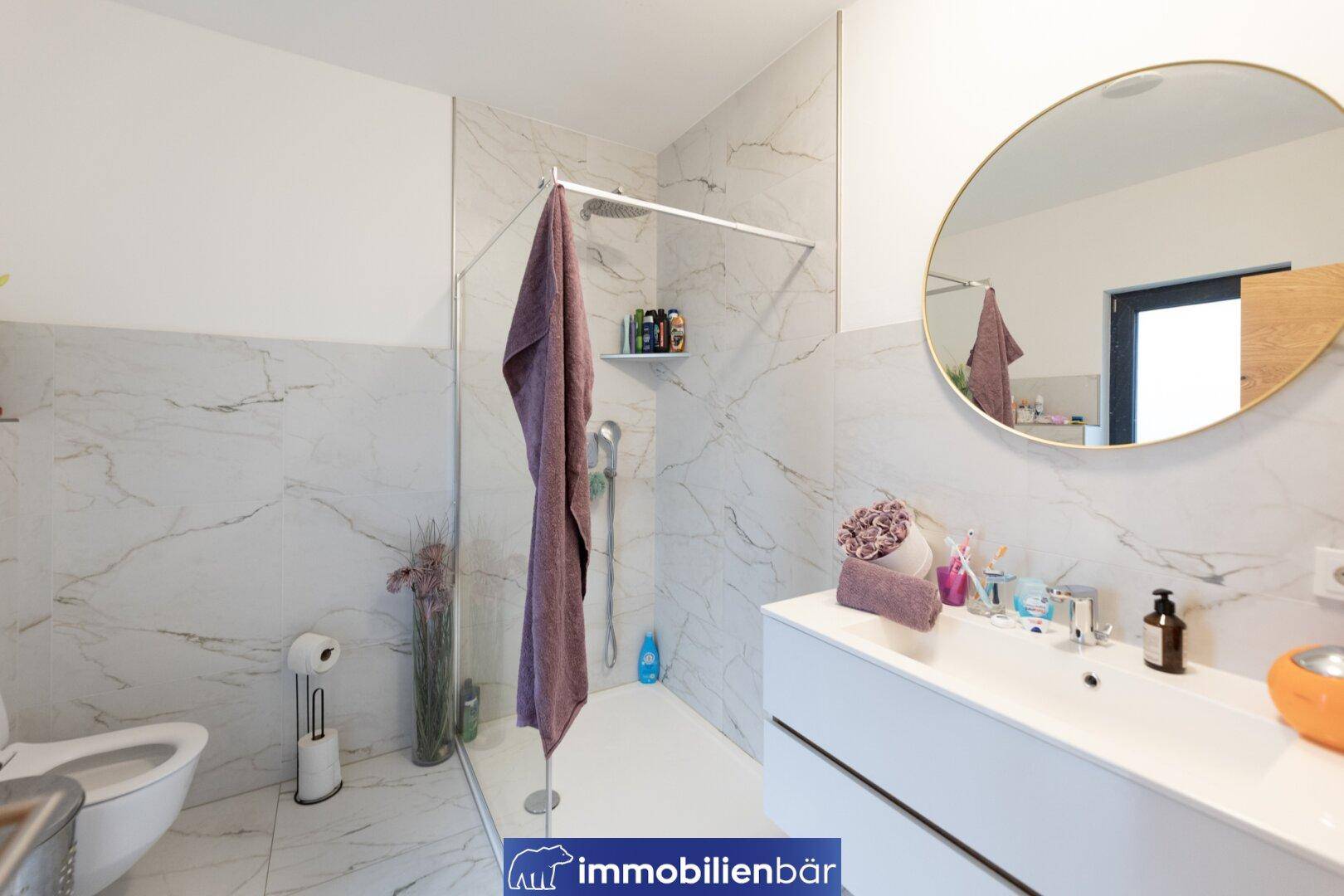 Modernes Badezimmer mit großformatigen Marmorfliesen, ebenerdiger Dusche und rundem Spiegel.