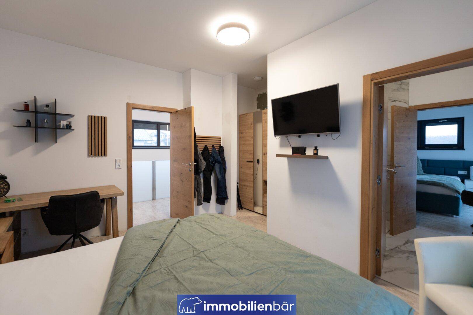 Helles Schlafzimmer mit Schreibtisch, Wandfernseher und Zugang zu weiteren Räumen.