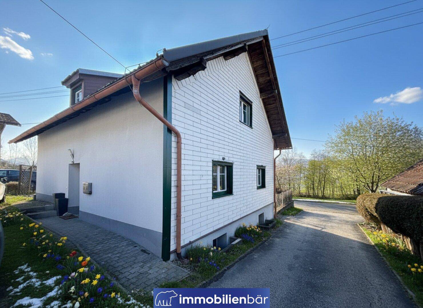 Seitenansicht des Hauses mit gepflastertem Weg, Blumenbeeten und ländlicher Umgebung.