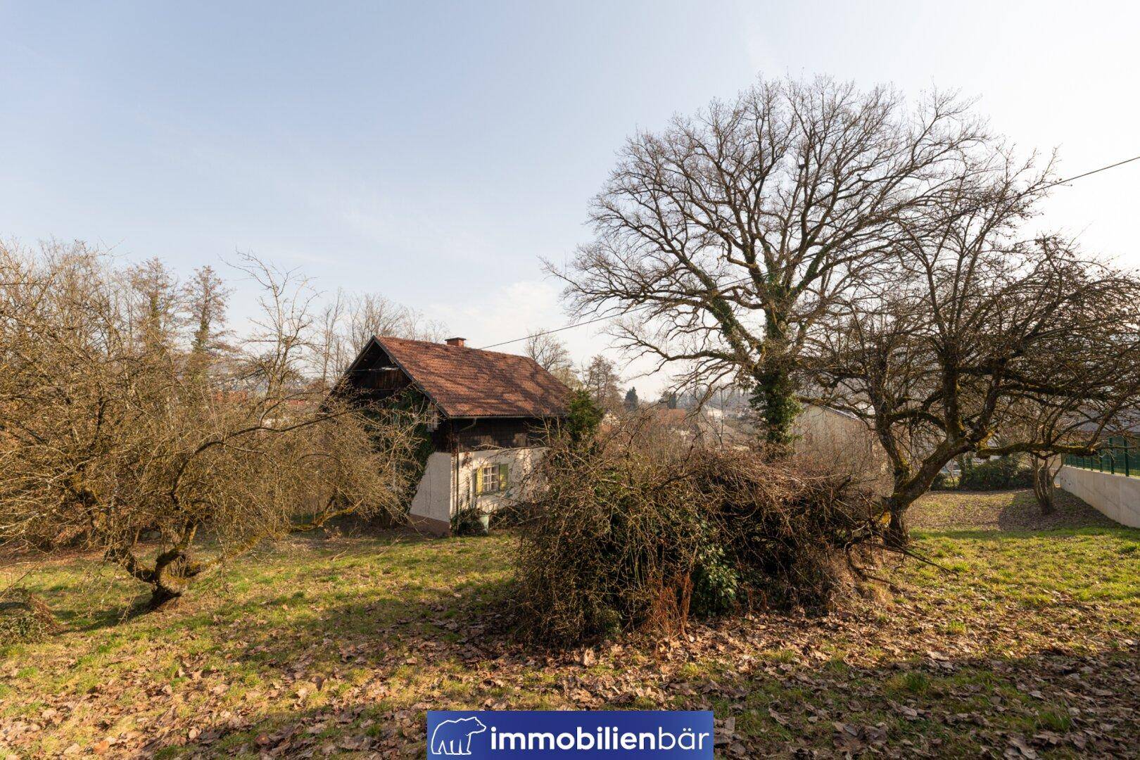 Altes Haus mit rotem Ziegeldach, umgeben von einem großen Garten und alten Bäumen.