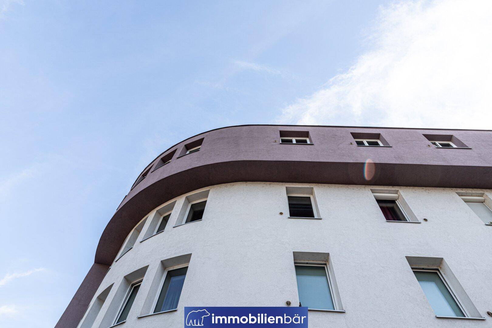 Moderne Fassade eines Mehrfamilienhauses mit abgerundeten Ecken und vielen Fenstern unter blauem Himmel.