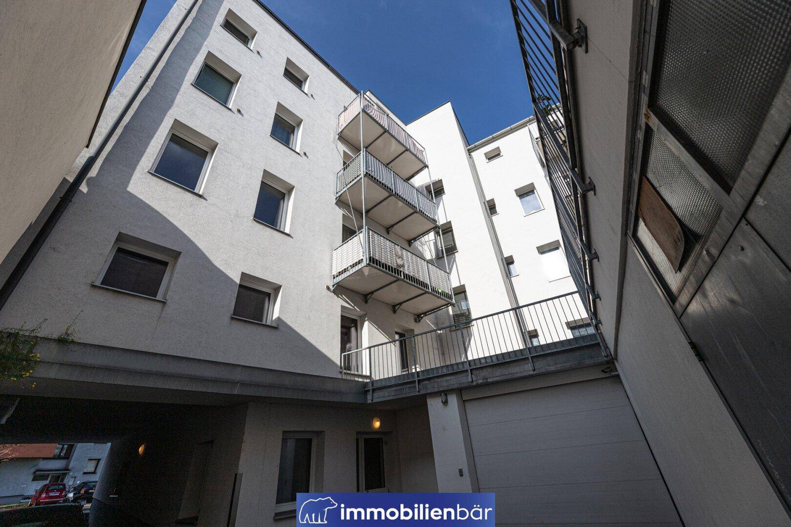 Innenhof eines Gebäudes mit mehreren Balkonen und einer Garageneinfahrt unter hellem Himmel.