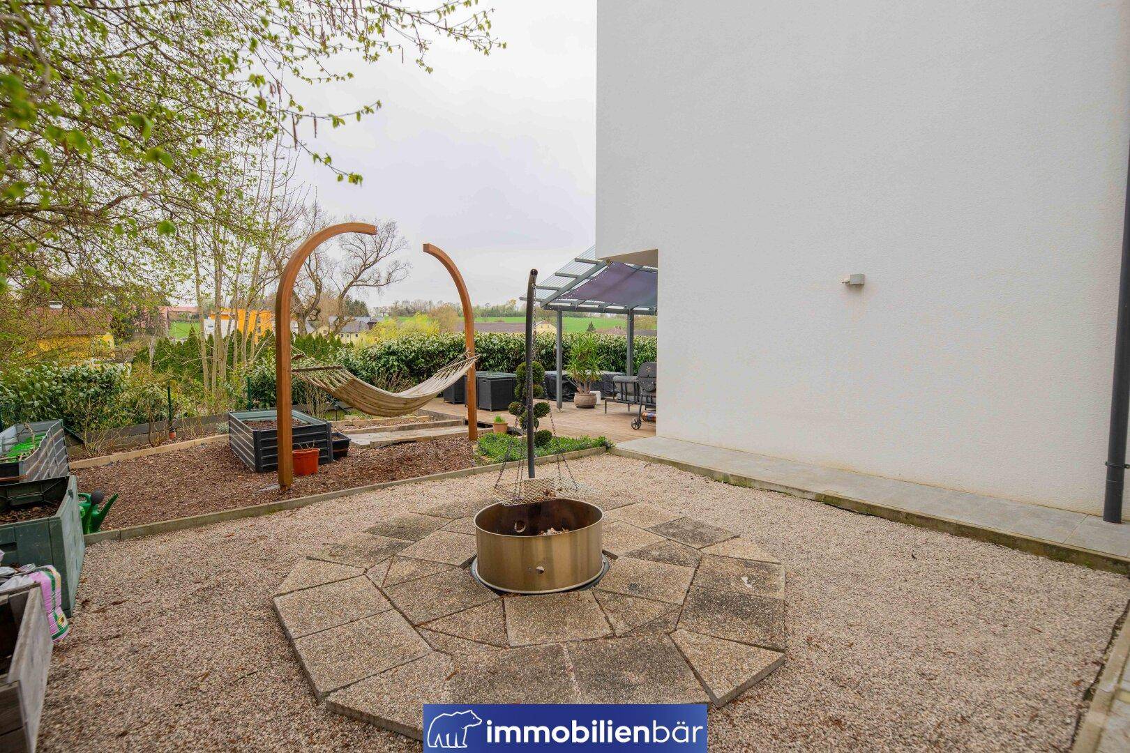 Gartenbereich mit Feuerstelle und Kiesfläche, umgeben von Grün und einer Hängematte.