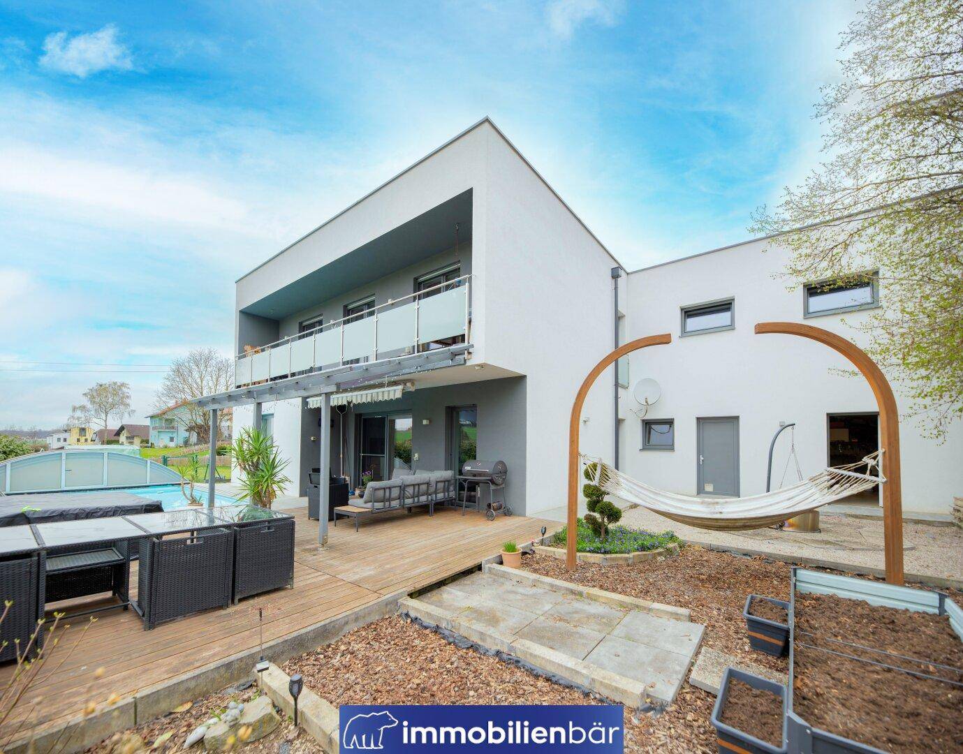 Moderne Villa mit großer Holzterrasse, Pool und Hängematte im Gartenbereich unter blauem Himmel.
